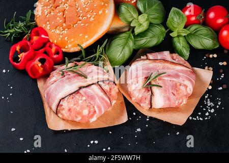 Two chicken hamburgers wrapped in bacon on cave paper with basil leaves, red peppers, cherry tomatoes and a hamburger bun, black background. Fast food Stock Photo
