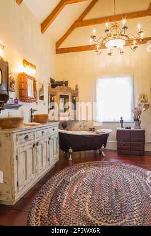 Bleached antique wooden general store cabinet with two molded bowl sinks and claw foot bathtub in main bathroom with weaved multicoloured oval rug Stock Photo