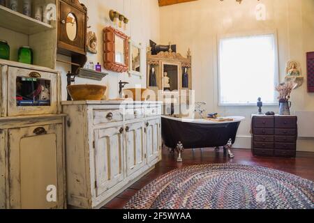 Bleached antique wooden general store cabinet with two molded bowl sinks and claw foot bathtub in main bathroom with weaved multicoloured oval rug Stock Photo