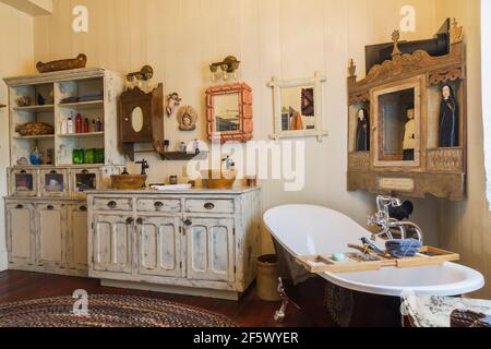 Bleached antique wooden general store cabinet with two molded bowl sinks and black claw foot bathtub in main bathroom with weaved multicoloured rug Stock Photo