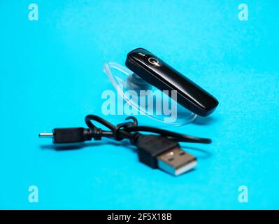 Simple hands-free wireless earpiece with a usb charging cable, object on a blue background, closeup, studio shot. Ergonomic mono handsfree headset wit Stock Photo