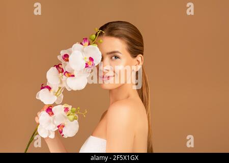 Pretty young lady covering eye with orchid, looking at camera and smiling, brown background. Spa, beauty and wellness Stock Photo
