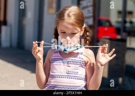 Young child, little girl putting on or taking off a colorful protective medical face mask, holding it in hands, portrait, closeup, outdoors shot, sunn Stock Photo