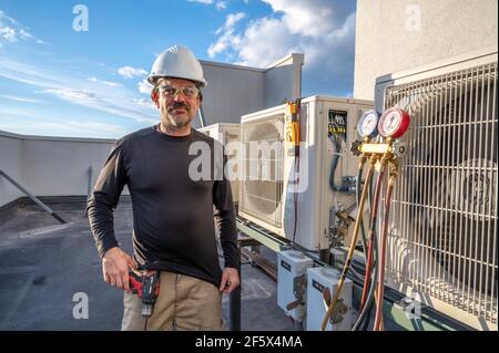 HVAC technician wearing safety gear standing next to mini-split ductless air conditioners on roof of building. Outdoors during the day. Stock Photo