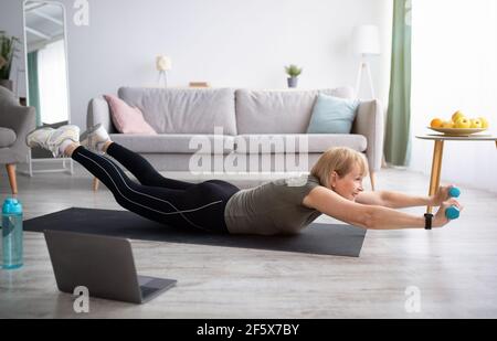 Strong mature woman making exercises with dumbbells on yoga mat, working out to video tutorial at home Stock Photo
