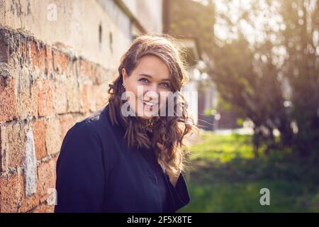 Happy smiling woman enjoying sunlight in city. Portrait of young caucasian woman with brown curly hair. Stock Photo