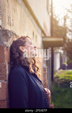 Happy brunette woman leaning brick wall and enjoying spring sunlight. Portrait of young brown hair woman Stock Photo