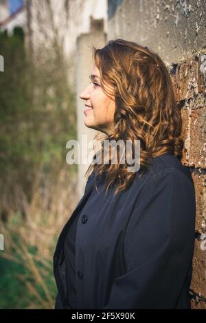 Attractive caucasian young woman with brown curly hair leaning brick wall. Portrait of smiling woman enjoying sunlight Stock Photo