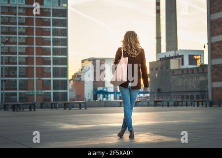 Fashionable blond hair woman is standing in city and enjoying sunset. Cityscape as background. Fashion and stylish concept Stock Photo