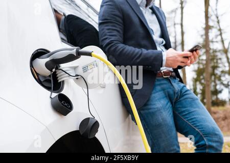 Close up charging an electric car and man standing near the vehicle and using his mobile phone.  Stock Photo