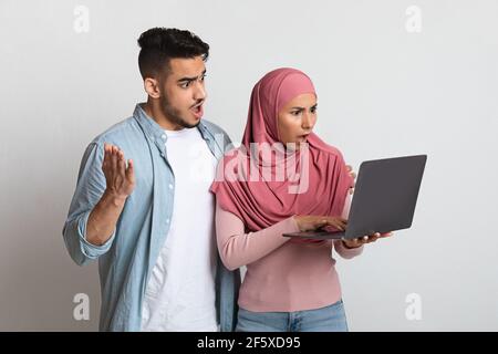 Shock Content. Surprised muslim couple holding laptop, emotionally reacting to internet news Stock Photo