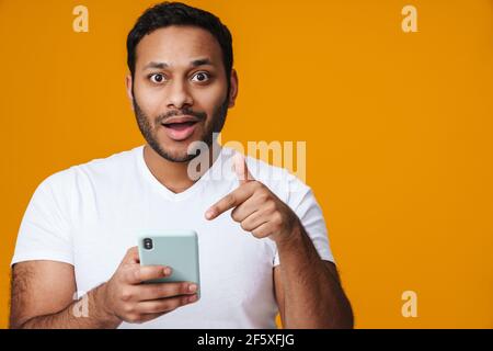 Asian surprised man holding and pointing finger at mobile phone isolated over yellow background Stock Photo