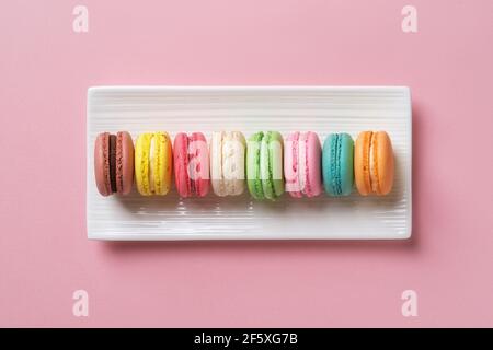 Multicolored macaroons on a white rectangular ceramic plate against pastel pink background. Sweet breakfast with delicious almond meringue cookies. Stock Photo