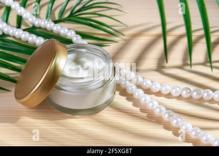 Anti-wrinkle moisturizing face cream in an open jar and pearl beads on a wooden desk. Green leaf casts a shadow on the hydrating facial gel glass jar. Stock Photo