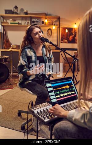 Young singer in headphones singing in microphone while woman using laptop to record her song in the studio Stock Photo