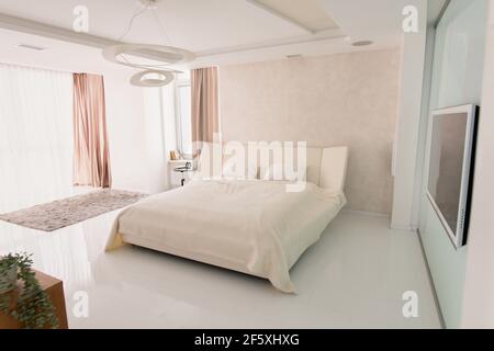 Interior of large cozy bedroom with comfortable double bed, plasma tv on wall, alarm clock on small table and grey furry rug on the floor Stock Photo