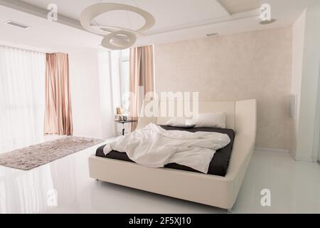 Interior of light bedroom with comfortable double bed, alarm clock on small table and grey furry rug on the floor and beige curtains on windows Stock Photo