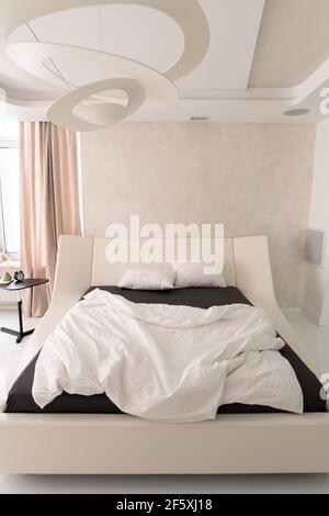 Interior of cozy bedroom with comfortable double bed with geometric chandelier above, alarm clock on small table and beige curtain on window Stock Photo