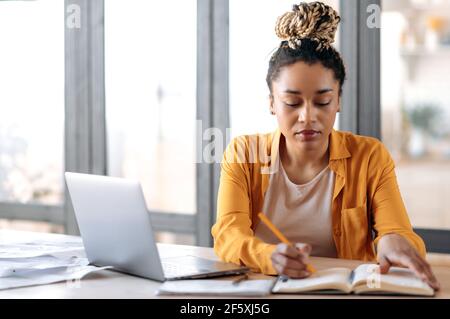 Focused smart stylish african american girl with dreadlocks, female student, in casual clothes, studying at home uses a laptop, watching online lecture, taking notes in notebook, sits at living room Stock Photo