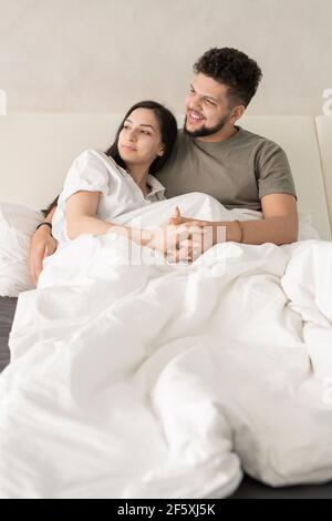 Young affectionate restful couple relaxing on comfortable bed in the morning and looking aside while discussing something pleasant Stock Photo