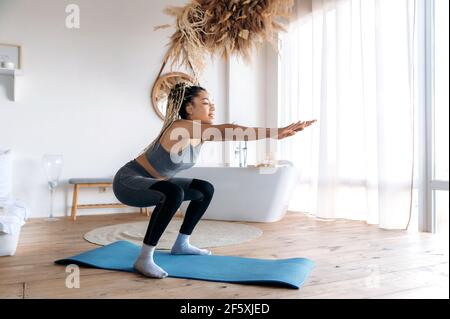 Healthy and sporty lifestyle. Attractive young African American slim sporty girl with dreadlocks care her health, does sports at home on fitness mat, does sit-ups with her arms outstretched, smiles Stock Photo