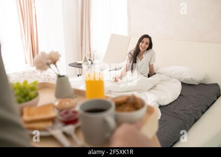 Happy young brunette female sitting under white blanket on large double bed and looking at her husband carrying tray with breakfast Stock Photo