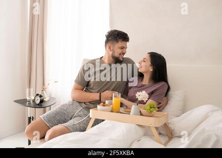 Happy young affectionate couple looking at one another with smiles while sitting in bed, having tasty breakfast and discussing something nice Stock Photo