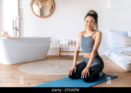 Attractive african american young sports woman with dreadlocks in sportswear, leads healthy lifestyle, sits at home on a fitness mat, ready for home sports workout, looks at the camera, smile friendly Stock Photo