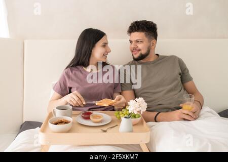 Happy young affectionate couple in t-shirts sitting in bed and talking during breakfast while female spreading peanut butter on toast Stock Photo