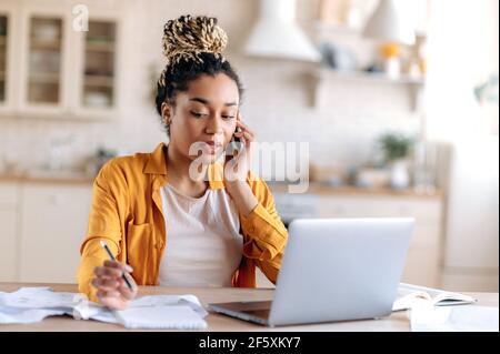 Confident successful busy African American woman with dreadlocks, manager or freelancer, working at home using a laptop, talking on a cell phone with a client, looking at notebook, taking notes Stock Photo