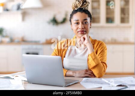 A serious intelligent young african american woman with dreadlocks, manager, freelancer or real estate agent in casual clothes, sits at the work desk, working from home, looks at camera