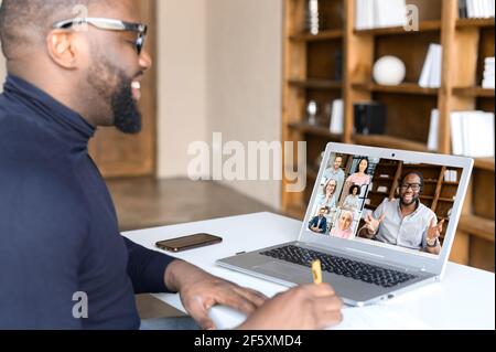 Male businessman with glasses and beard leads distant communication with multicultural colleagues using laptop app, discussing world social project, writing down plans and ideas, sitting at home Stock Photo