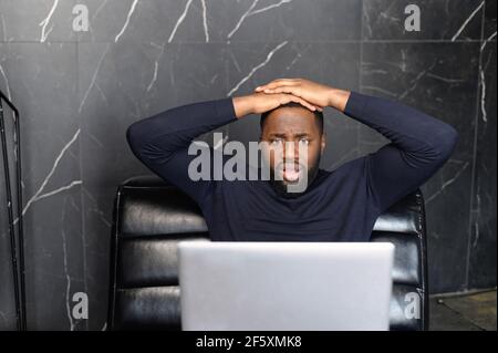 Oh no. Portrait of shocked black businessman holding hands on head, looking very disappointed, looking at screen of computer eyes wide open, thinking received bad news, waiting hoping positive result Stock Photo
