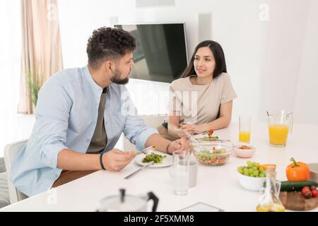 Young brunette woman looking at her husband during conversation by breakfast while both having selfmade vegetable salad and orange juice Stock Photo