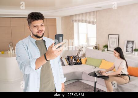 Young bearded man in casualwear holding smartphone in front of himself while talking to his friend against woman using laptop on sofa Stock Photo