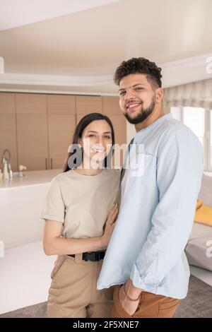 Young cheerful couple in casualwear looking at you with toothy smiles while standing close to one another in front of camera at home Stock Photo