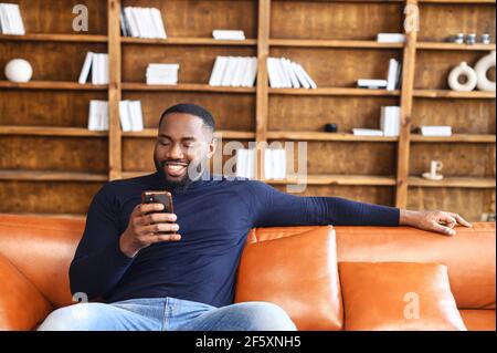 Portrait of smiling african american man sitting on sofa in office, using mobile phone for checking social nets, typing message for family, girlfriend, business partner, views photos, watches videos Stock Photo