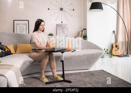 Young pretty female with dark long hair sitting on couch in front of small table with laptop and touching buttons of keypad in living-room Stock Photo