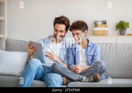 Joyful father and son using tablet computer, watching videos or playing online games, spending time together at home, Stock Photo