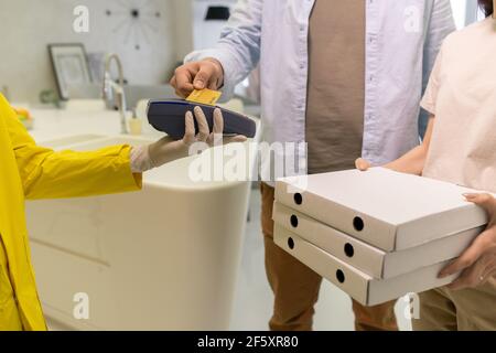 Young man in casualwear holding credit card over payment machine held by gloved courier while female holding stack of boxes with pizza Stock Photo