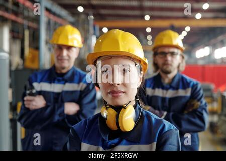 Young serious female worker of large industrial plant or factory in workwear and protective helmet standing against two male colleagues Stock Photo
