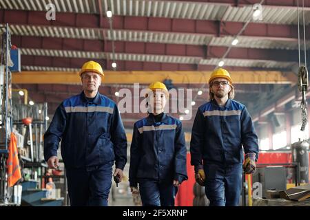 Group of successful male and female workers or engineers of contemporary factory moving along workshop with industrial equipment Stock Photo