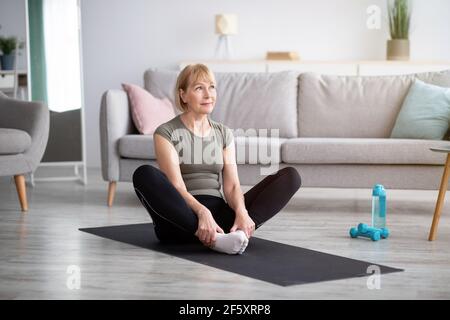 Full length of athletic mature woman stretching her legs, sitting in butterfly yoga pose on mat indoors, free space Stock Photo