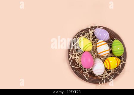 Color easter eggs in rustic plate with straw on a beige pastel background. Easter background. Festive food ornament. Greeting card. Stock Photo
