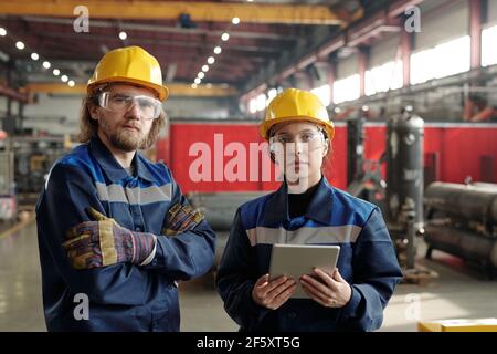 Two young serious engineers in protective workwear looking at you while standing in front of camera, female worker using digital tablet Stock Photo