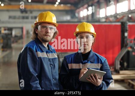 Two young serious engineers in protective hardhats and workwear looking at you while standing in front of camera inside large workshop Stock Photo
