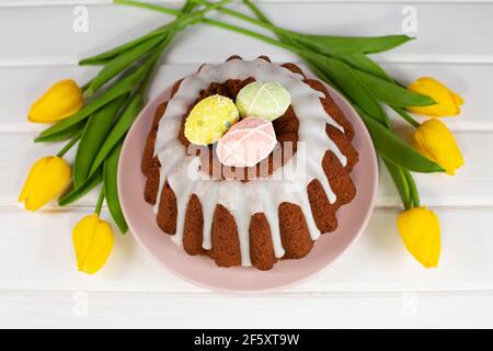 Glazed easter cake decorated with mini eggs candy and yellow tulips on wooden table. Happy Easter Holidays. Stock Photo