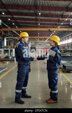 Two young successful engineers in protective hardhats and workwear looking at one another while handshaking inside large workshop Stock Photo