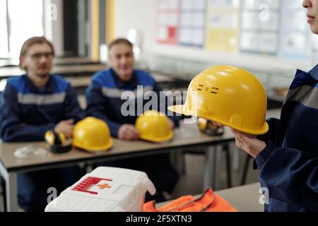 Young female worker in uniform holding protective helmet while standing in front of male colleagues at the lesson of basics of life safety Stock Photo