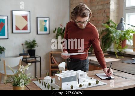Young serious architect in casualwear and eyeglasses making notes while standing by table with model of new house and yard in office Stock Photo
